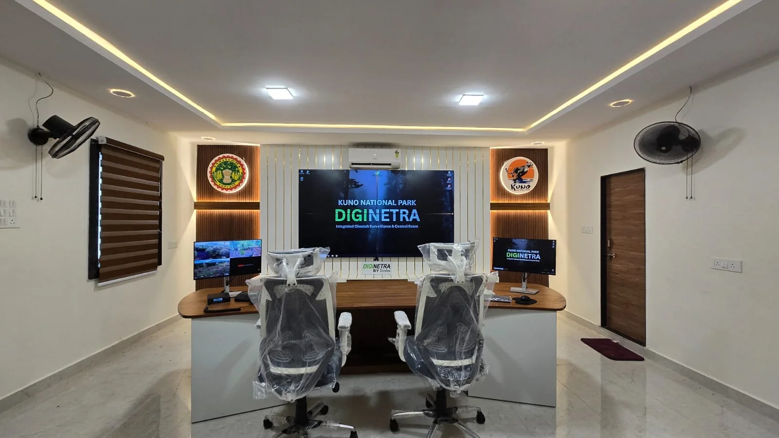 The DigiNetra control room inside Kuno National Park: two operator workstations facing a wall-mounted display reading “Kuno National Park DigiNetra — Integrated Cheetah Surveillance & Control Room”, with live camera feeds on a side monitor.